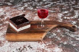 A slice of layered chocolate cake with a glass of red wine on a wooden serving board.