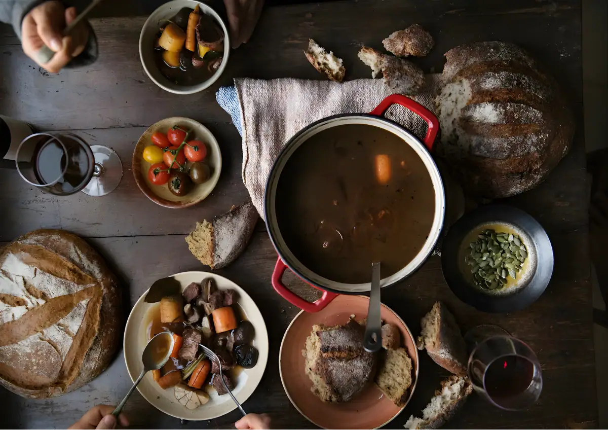 Cozy rustic meal with bread, stew, and fresh vegetables on a wooden table.